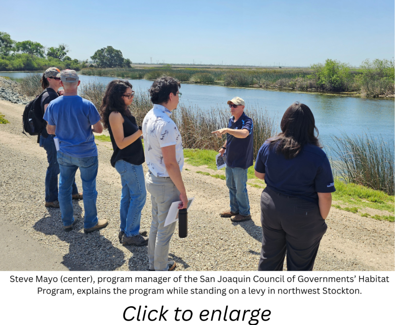 Group of people on a levy near water and farm fields talking. A caption and \